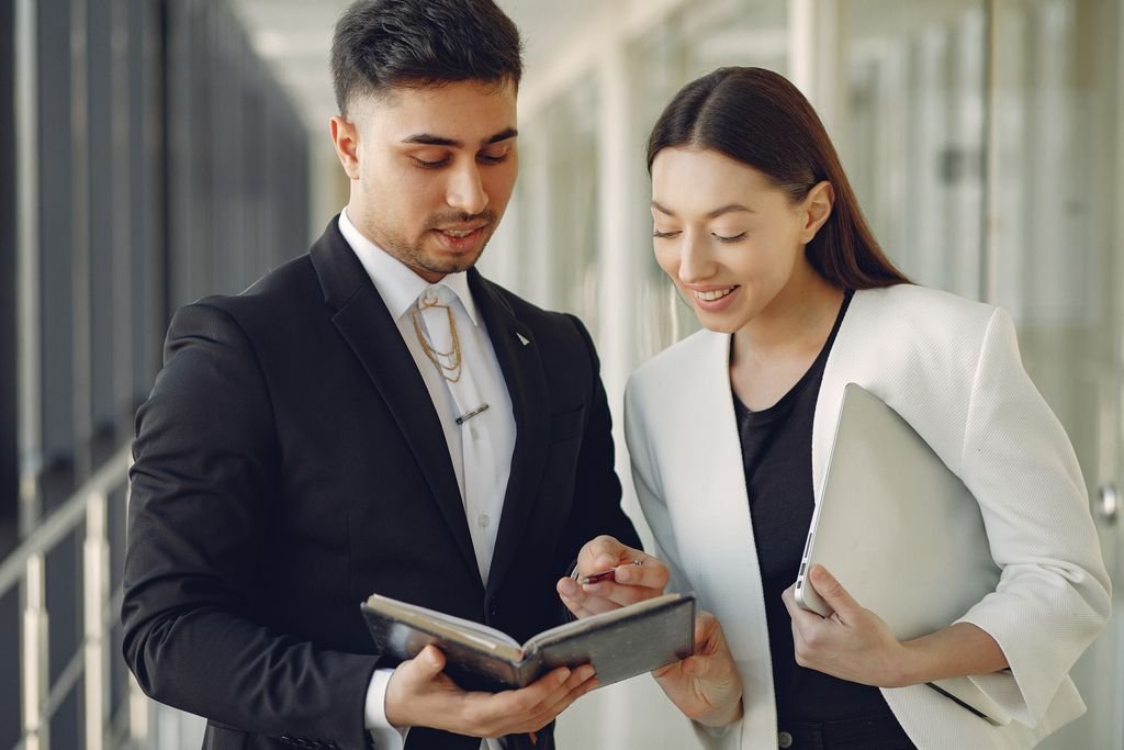 Focused diverse coworkers in formal clothes with notebook and laptop in hands standing in hall and discussing business project together
